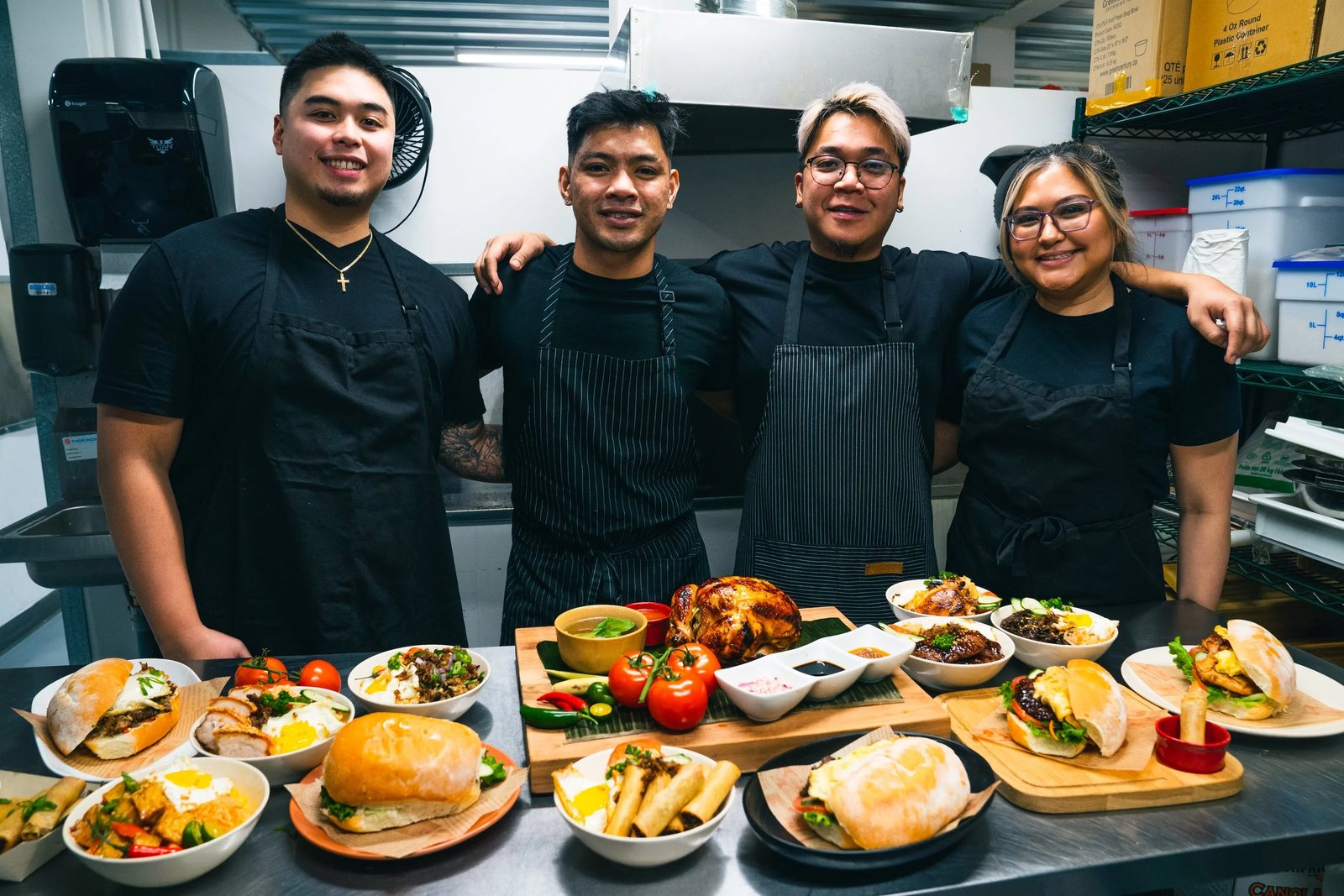 The four friends behind Halina in black aprons in the kitchen, behind a table of plated Filipino dishes